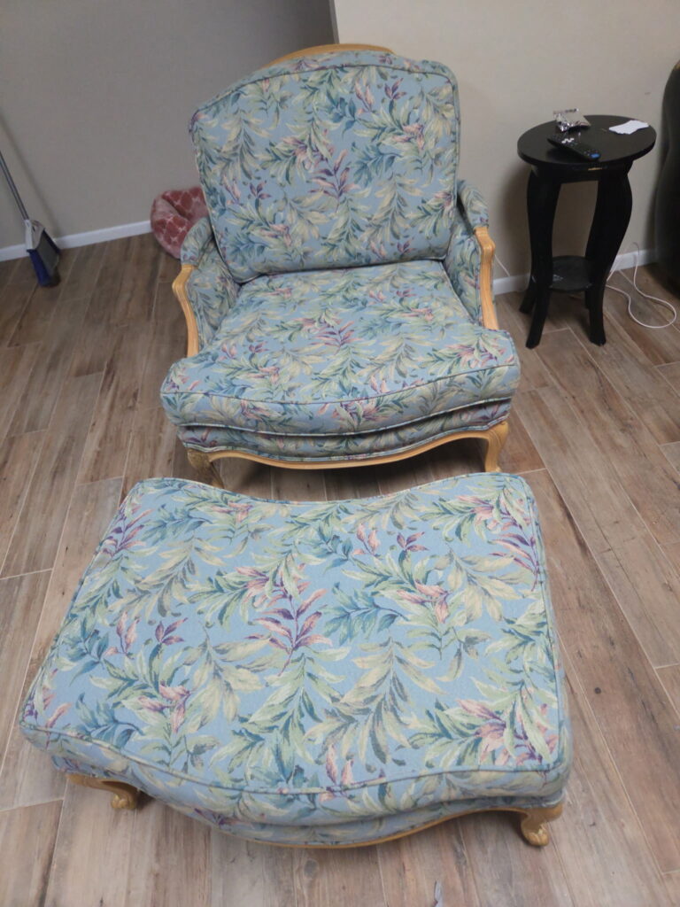 Front view of a vintage wood-framed accent chair and matching large ottoman upholstered in a dated pastel blue, green, and pink botanical leaf fabric.